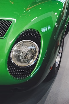 A close-up view of a green vintage car's front headlight and part of the fender. The paint is glossy, reflecting the surrounding lights and creating highlights on the curved surface. The headlight is circular and surrounded by a ribbed, black trim.