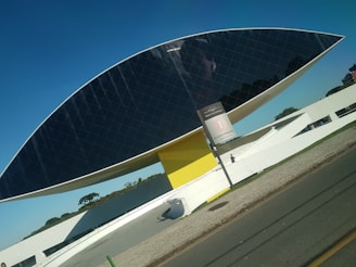 A modern architectural structure resembling an eye, supported by a yellow pillar and featuring reflective dark glass panels. It is set against a clear blue sky with a pedestrian walking nearby on a paved pathway. A sign indicating 'Acesso Principal' is visible in the foreground.