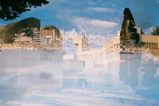 A double exposure image combining urban architecture with a natural landscape, featuring buildings interwoven with sky and trees. The result is a surreal blending of geometric and organic forms, creating an abstract composition.