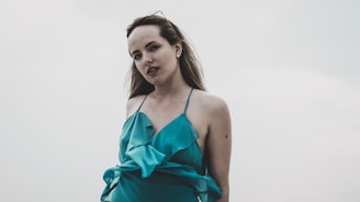 A confident woman wearing a soft pastel dress, standing calmly in a minimalist, sunlit room.