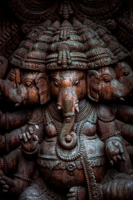 A detailed wooden sculpture featuring multiple faces of an elephant-headed deity, adorned with intricate carvings and ornate headgear. The craftsmanship highlights elaborate patterns and textures.