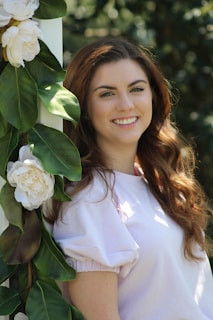 A young woman with long, wavy brown hair is smiling. She is wearing a light-colored top and is leaning against a structure adorned with white flowers and green leaves. The background is a blurred green setting, suggesting an outdoor, natural environment.