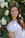 A young woman with long, wavy brown hair is smiling. She is wearing a light-colored top and is leaning against a structure adorned with white flowers and green leaves. The background is a blurred green setting, suggesting an outdoor, natural environment.