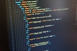 A computer monitor displays HTML code in an integrated development environment. The screen shows a structured hierarchy of HTML tags, including elements such as header, nav, and list items with classes and hyperlinks.