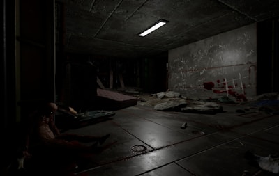 A dimly lit, sinister room with a concrete ceiling and floor, featuring scattered debris and a mattress. Graffiti and red marks resembling bloodstains cover one wall. A humanoid figure slumps against a wall, adding to the eerie atmosphere, while a flickering overhead light casts shadows.