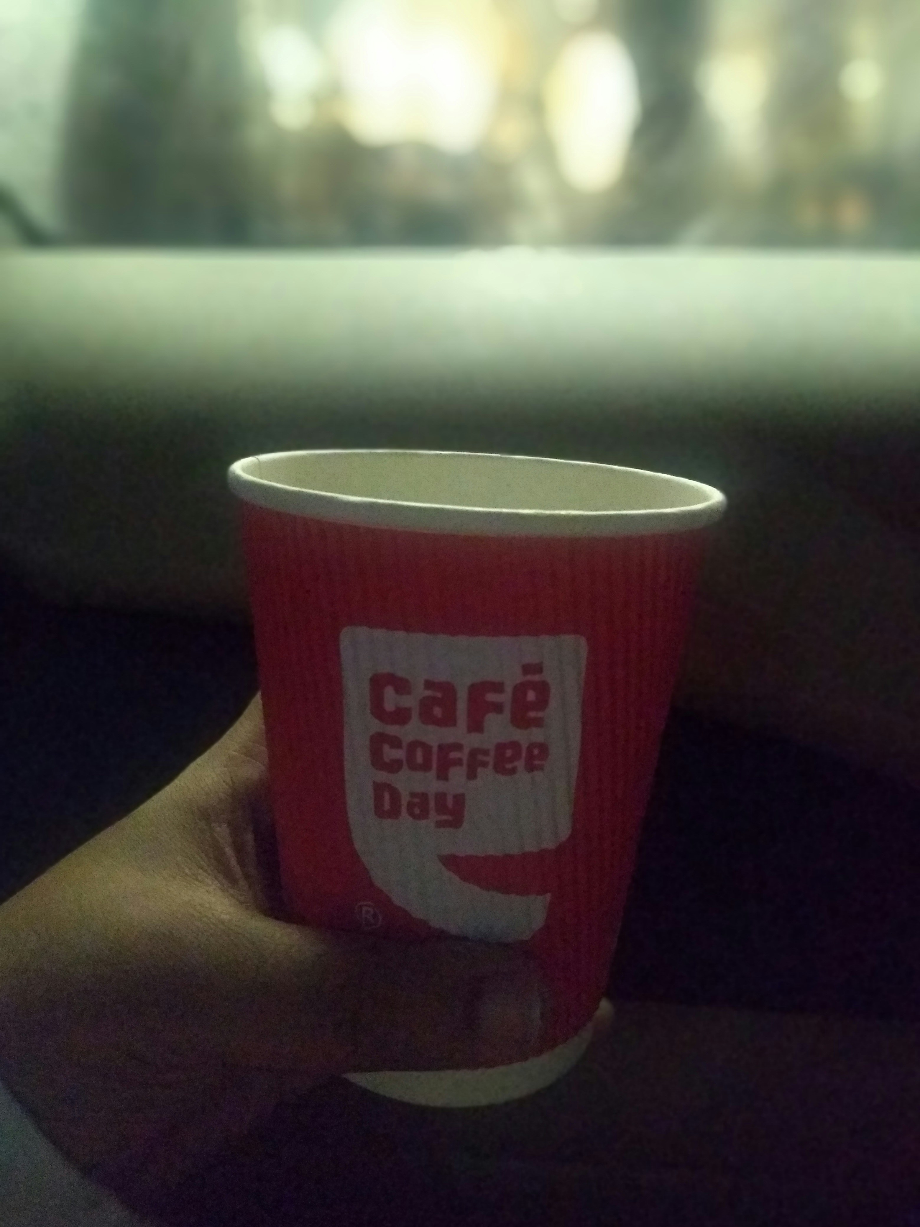 A hand holds a red paper cup with the logo and text 'Café Coffee Day' against a blurred background. The cup is ribbed, and the lighting suggests a cozy, dimly lit setting.