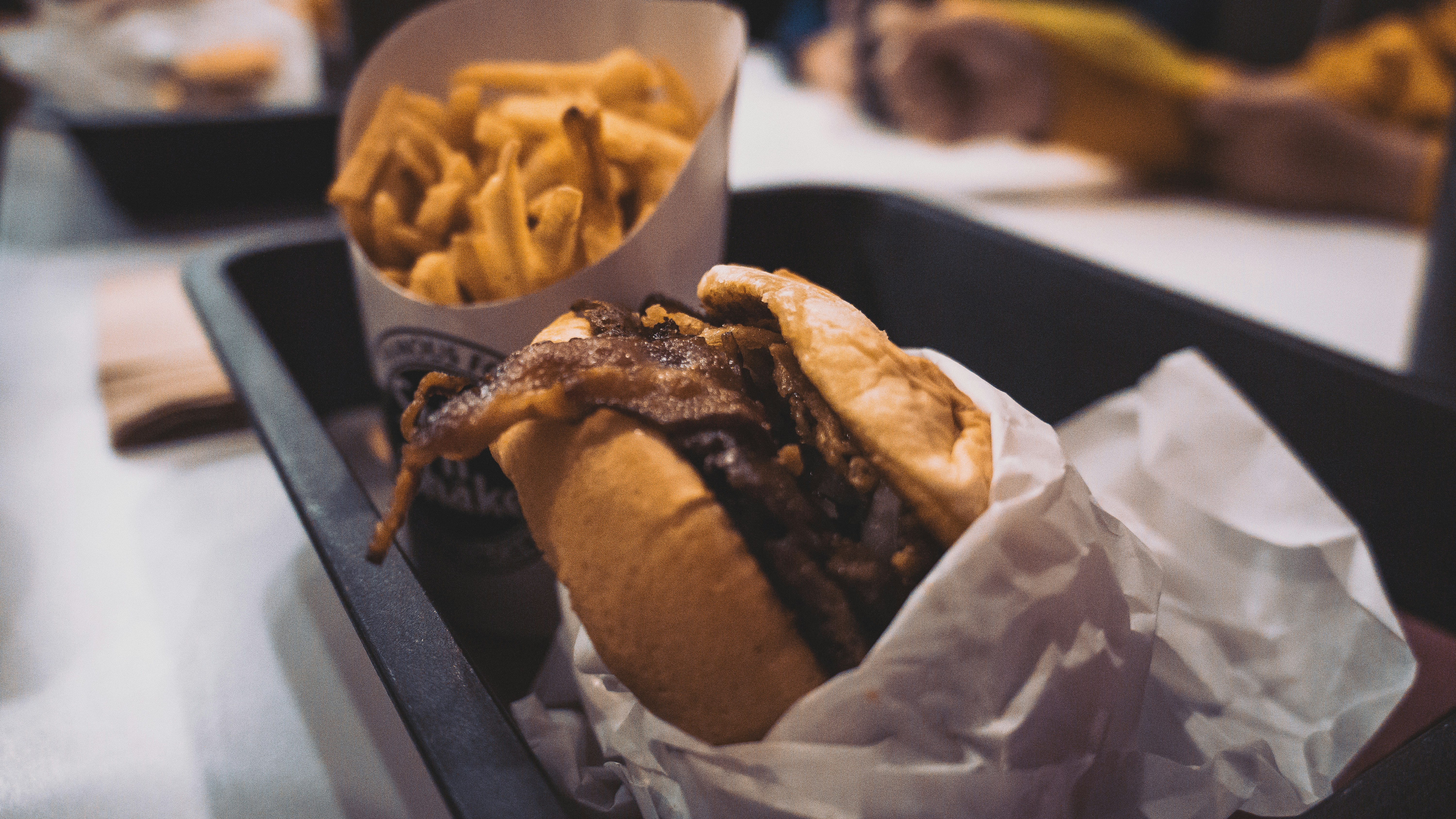 A close-up of a tray containing a burger with crispy onions and a side of golden French fries. The burger is wrapped in white paper, and the tray is set on a table in what appears to be a casual dining setting.