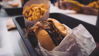 A close-up of a tray containing a burger with crispy onions and a side of golden French fries. The burger is wrapped in white paper, and the tray is set on a table in what appears to be a casual dining setting.