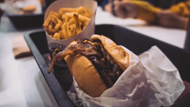 A close-up of a tray containing a burger with crispy onions and a side of golden French fries. The burger is wrapped in white paper, and the tray is set on a table in what appears to be a casual dining setting.