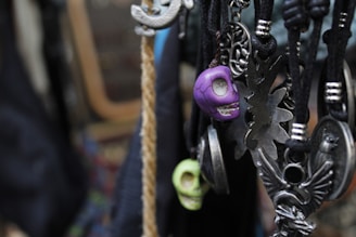 Various pendants and charms hang on black cords, including skull-shaped and ornate metallic designs. The focus is on a purple skull pendant, surrounded by intricate metallic figures and shapes.