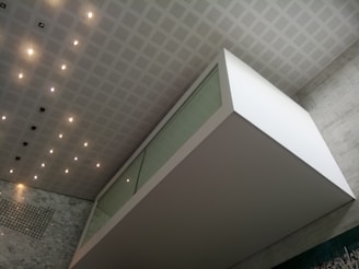 An architectural interior featuring a modern, geometric design with a floating glass and white structure against a wall of gray marble. The ceiling is patterned with a grid of recessed lights and panels, enhancing the clean and contemporary atmosphere.