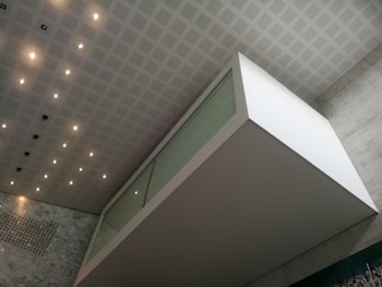 An architectural interior featuring a modern, geometric design with a floating glass and white structure against a wall of gray marble. The ceiling is patterned with a grid of recessed lights and panels, enhancing the clean and contemporary atmosphere.
