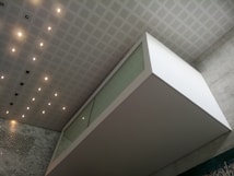 An architectural interior featuring a modern, geometric design with a floating glass and white structure against a wall of gray marble. The ceiling is patterned with a grid of recessed lights and panels, enhancing the clean and contemporary atmosphere.
