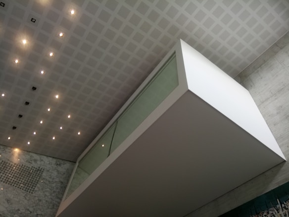 An architectural interior featuring a modern, geometric design with a floating glass and white structure against a wall of gray marble. The ceiling is patterned with a grid of recessed lights and panels, enhancing the clean and contemporary atmosphere.