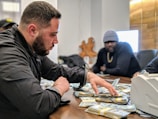 A man is sorting through a large pile of cash on a table, with another man sitting behind him. They appear to be in a dimly lit office setting. The man in the foreground is focused, wearing a black jacket. The mood is tense and serious.