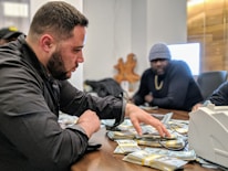 A man is sorting through a large pile of cash on a table, with another man sitting behind him. They appear to be in a dimly lit office setting. The man in the foreground is focused, wearing a black jacket. The mood is tense and serious.