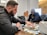 A man is sorting through a large pile of cash on a table, with another man sitting behind him. They appear to be in a dimly lit office setting. The man in the foreground is focused, wearing a black jacket. The mood is tense and serious.