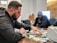 A man is sorting through a large pile of cash on a table, with another man sitting behind him. They appear to be in a dimly lit office setting. The man in the foreground is focused, wearing a black jacket. The mood is tense and serious.