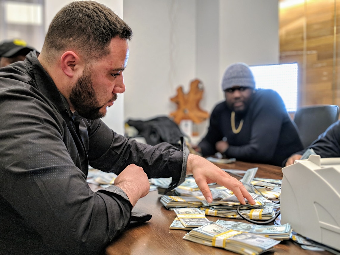 A man is sorting through a large pile of cash on a table, with another man sitting behind him. They appear to be in a dimly lit office setting. The man in the foreground is focused, wearing a black jacket. The mood is tense and serious.