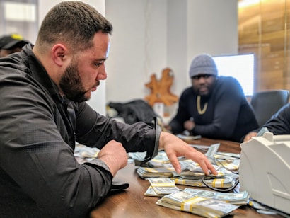 A man is sorting through a large pile of cash on a table, with another man sitting behind him. They appear to be in a dimly lit office setting. The man in the foreground is focused, wearing a black jacket. The mood is tense and serious.