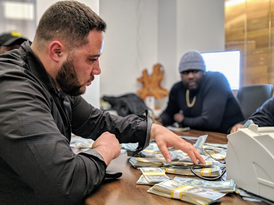 A man is sorting through a large pile of cash on a table, with another man sitting behind him. They appear to be in a dimly lit office setting. The man in the foreground is focused, wearing a black jacket. The mood is tense and serious.