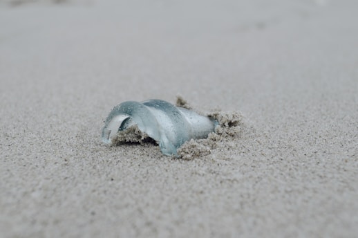 A broken piece of glass is partially buried in the sand, surrounded by fine grains. The smooth curvature and transparency of the glass contrast with the rough texture of the sand.