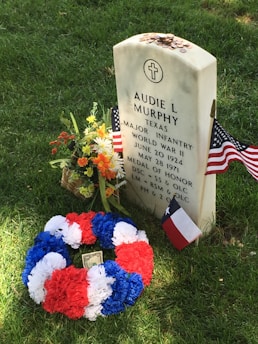 A gravestone with white and blue engraved text, surrounded by United States and Texas flags. A colorful flower wreath in red, white, and blue is placed at the base, along with a bouquet of fresh flowers. Coins are scattered on top of the gravestone, which is set on a grassy area.