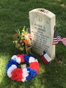 A gravestone with white and blue engraved text, surrounded by United States and Texas flags. A colorful flower wreath in red, white, and blue is placed at the base, along with a bouquet of fresh flowers. Coins are scattered on top of the gravestone, which is set on a grassy area.
