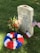 A gravestone with white and blue engraved text, surrounded by United States and Texas flags. A colorful flower wreath in red, white, and blue is placed at the base, along with a bouquet of fresh flowers. Coins are scattered on top of the gravestone, which is set on a grassy area.