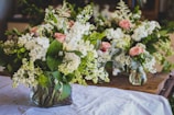 A collection of floral arrangements with white and pink flowers, including roses and various greenery, displayed in transparent vases on a table with a white cloth. The scene suggests a fresh and elegant atmosphere.