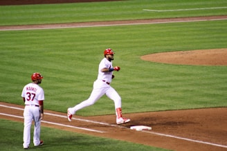 A baseball player in a white and red uniform is running towards a base on a well-maintained field. Another player, also in a white and red uniform with the number 37, stands nearby, observing. The game takes place on a brightly lit, green grass field with brown dirt areas around the bases.