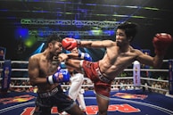 Two athletes are engaged in a dynamic kickboxing match inside a ring, surrounded by a cheering audience. One fighter, wearing red gloves and shorts, executes a high kick towards his opponent, who attempts to block the attack with his arm. The background features colorful lighting and promotional banners.