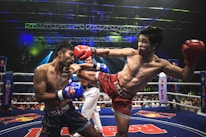 Two athletes are engaged in a dynamic kickboxing match inside a ring, surrounded by a cheering audience. One fighter, wearing red gloves and shorts, executes a high kick towards his opponent, who attempts to block the attack with his arm. The background features colorful lighting and promotional banners.
