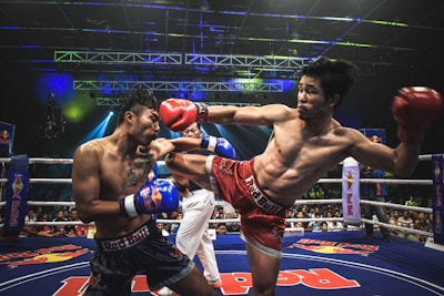 Two athletes are engaged in a dynamic kickboxing match inside a ring, surrounded by a cheering audience. One fighter, wearing red gloves and shorts, executes a high kick towards his opponent, who attempts to block the attack with his arm. The background features colorful lighting and promotional banners.