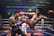 Two athletes are engaged in a dynamic kickboxing match inside a ring, surrounded by a cheering audience. One fighter, wearing red gloves and shorts, executes a high kick towards his opponent, who attempts to block the attack with his arm. The background features colorful lighting and promotional banners.