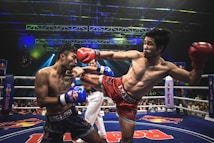 Two athletes are engaged in a dynamic kickboxing match inside a ring, surrounded by a cheering audience. One fighter, wearing red gloves and shorts, executes a high kick towards his opponent, who attempts to block the attack with his arm. The background features colorful lighting and promotional banners.