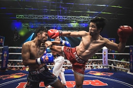 Two athletes are engaged in a dynamic kickboxing match inside a ring, surrounded by a cheering audience. One fighter, wearing red gloves and shorts, executes a high kick towards his opponent, who attempts to block the attack with his arm. The background features colorful lighting and promotional banners.