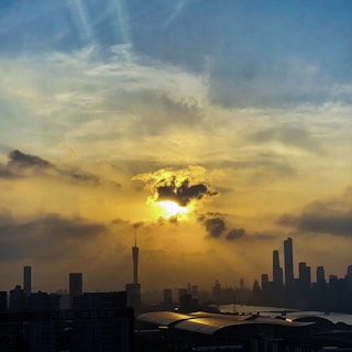 The image captures a cityscape at sunset with a dramatic sky. The sun is partially obscured by clouds, casting a warm, golden light over the skyline. Tall buildings, including a distinctive tower, are silhouetted against the vibrant sky. The river reflects the sunset's glow, adding to the scene's beauty.