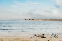 A serene beach scene with two bicycles parked on the sand and a couple sitting close to each other, facing the ocean. The water is calm, with gentle waves lapping the shore. In the distance, a few people are swimming. The sky is partly cloudy, with hues of blue and white.
