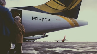A group of people is boarding a plane on an empty tarmac. The tail of the aircraft is prominently displayed with a design featuring yellow and black colors. In the background, another airplane is parked on the tarmac under an overcast sky.