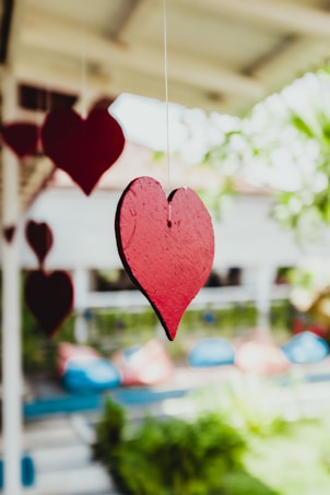 Several red heart-shaped decorations hang from strings against a blurred background of greenery and colorful beanbags.