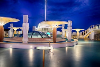 A modern, well-lit cruise ship deck at night with futuristic architectural structures. The deck features glass domes, round tables, and artistic lighting fixtures. The deep blue evening sky enhances the ambiance.