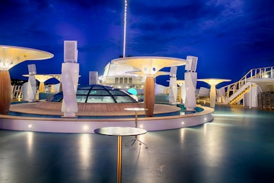 A modern, well-lit cruise ship deck at night with futuristic architectural structures. The deck features glass domes, round tables, and artistic lighting fixtures. The deep blue evening sky enhances the ambiance.