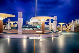 A modern, well-lit cruise ship deck at night with futuristic architectural structures. The deck features glass domes, round tables, and artistic lighting fixtures. The deep blue evening sky enhances the ambiance.