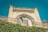 An ornate, vintage architectural facade with intricate carvings and details is prominently displayed, topped by a decorative archway with a broken arched window. The structure features a weathered stone facade and a wrought iron balcony railing adorned with ivy, set against a clear blue sky.