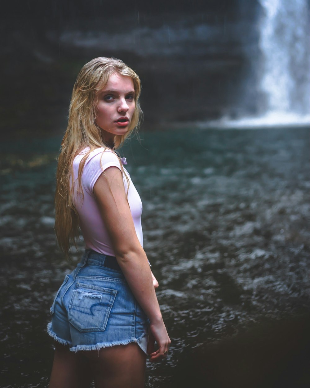 selective focus photography of woman wearing pink shirt and blue denim short shorts standing on front of waterfalls