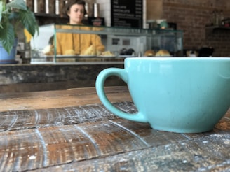 A turquoise coffee cup sits on a rustic wooden table. In the background, there is a display case with pastries and a person standing behind it, partially out of focus. The setting appears to be a cozy cafe with brick walls and a casual atmosphere.