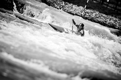 A kayaker navigates through turbulent white-water rapids, captured in a dramatic black and white photograph. The water splashes intensely around the athlete, who is focused, wearing a helmet and holding a paddle.