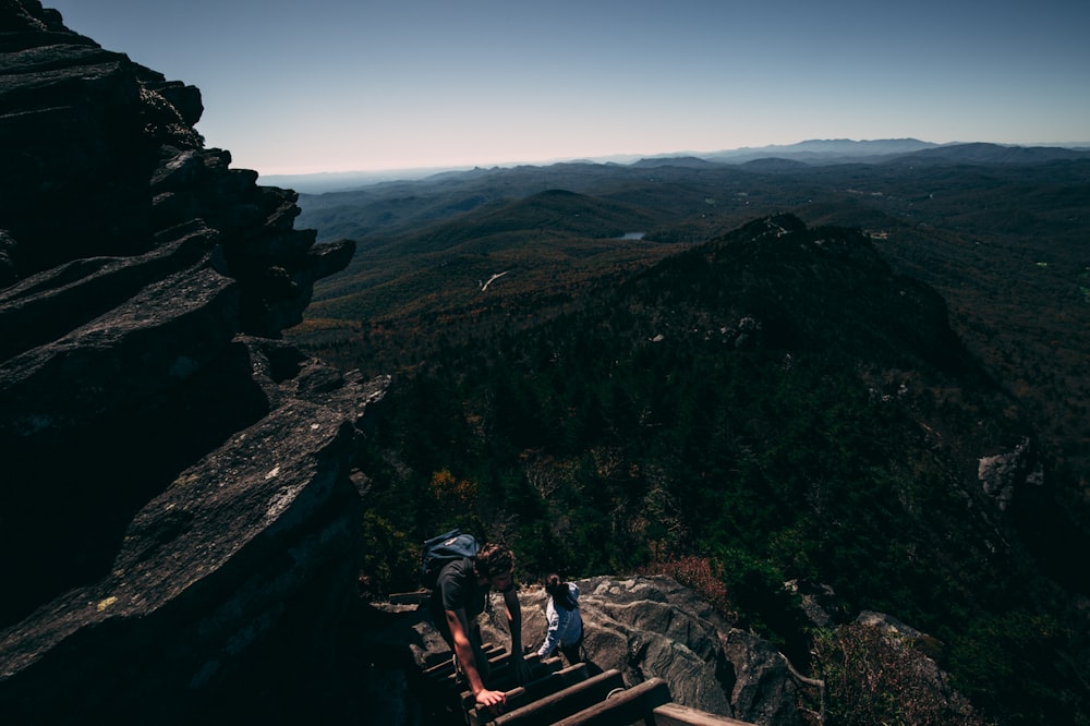 2 men sitting on brown wooden bench on top of mountain during daytime