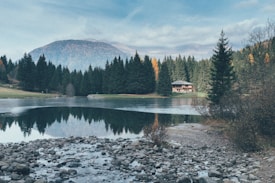 A serene landscape featuring a clear, reflective lake surrounded by dense pine forests. A small cabin can be seen nestled among the trees on the right side of the image. In the background, a mountain peak rises, partially covered by mist. The foreground contains a rocky shoreline with scattered stones, giving a natural and rustic feel.